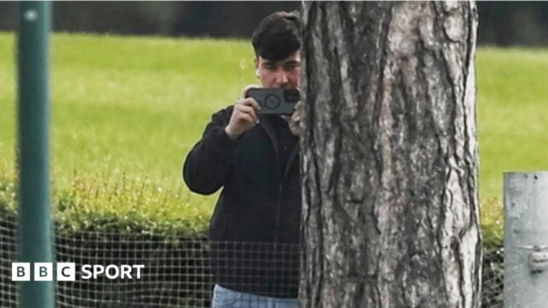 Southampton expelled from play-offs for spying on Middlesbrough, Oxford and Ipswich A man in a dark jacket and blue jeans stands partially concealed by a tree trunk and holds a phone up in two hands as if filming and has a white earphone in his right ear