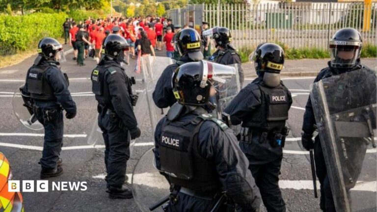 Seven police officers are in riot gear. They have helmets on and their faces are covered in a plastic shield. Football fans wearing red tops are in the background.