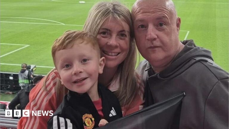A family of 3 stand in a stadium, which is filling up, smiling to a camera on matchday.