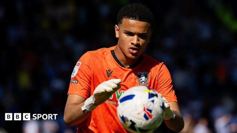 Pierce Charles concentrates on catching the ball in his hands, wearing orange goalkeeping jersey